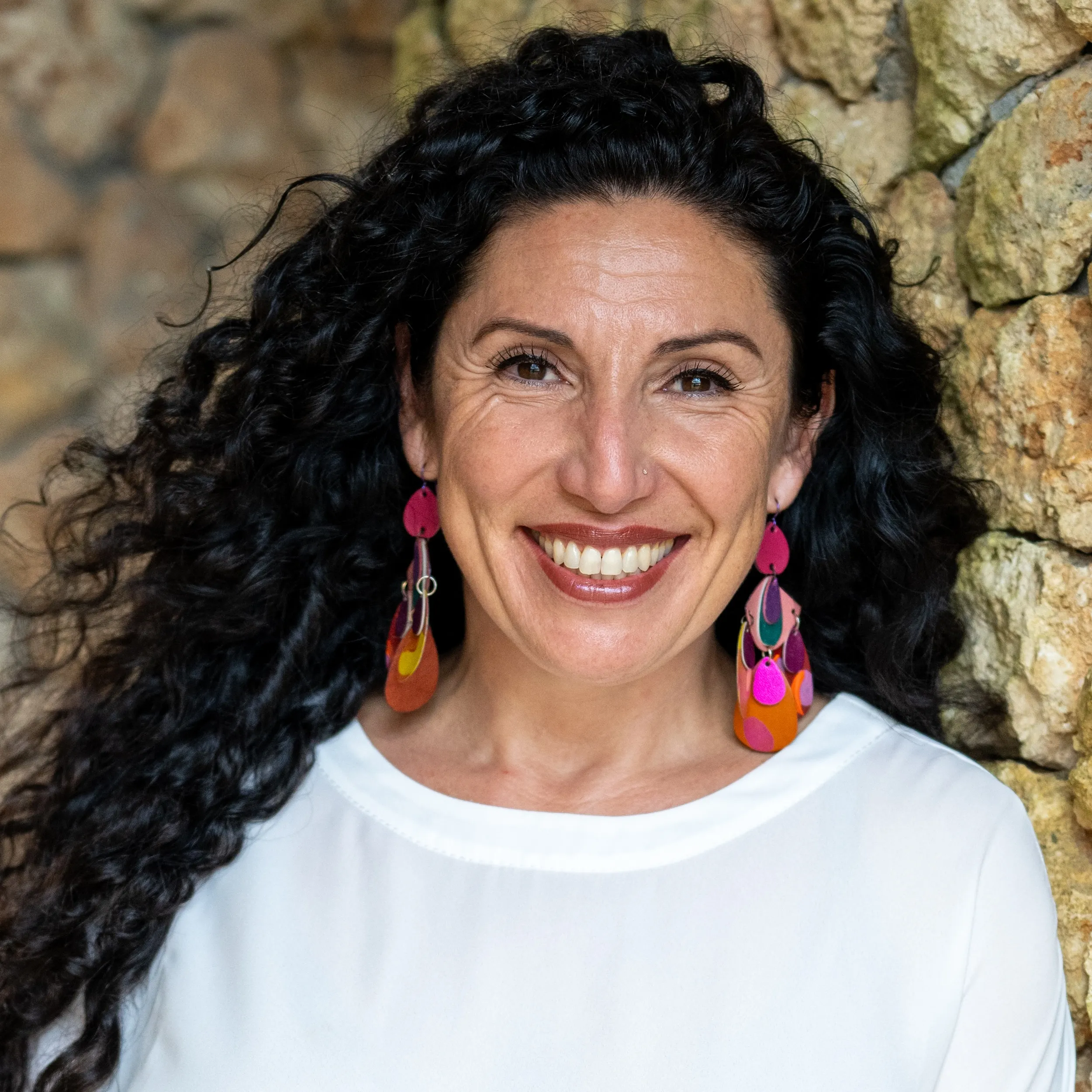 Close-up photo of Maritza Schafer wearing a white top, colorful earrings, and leaning against a textured stone wall