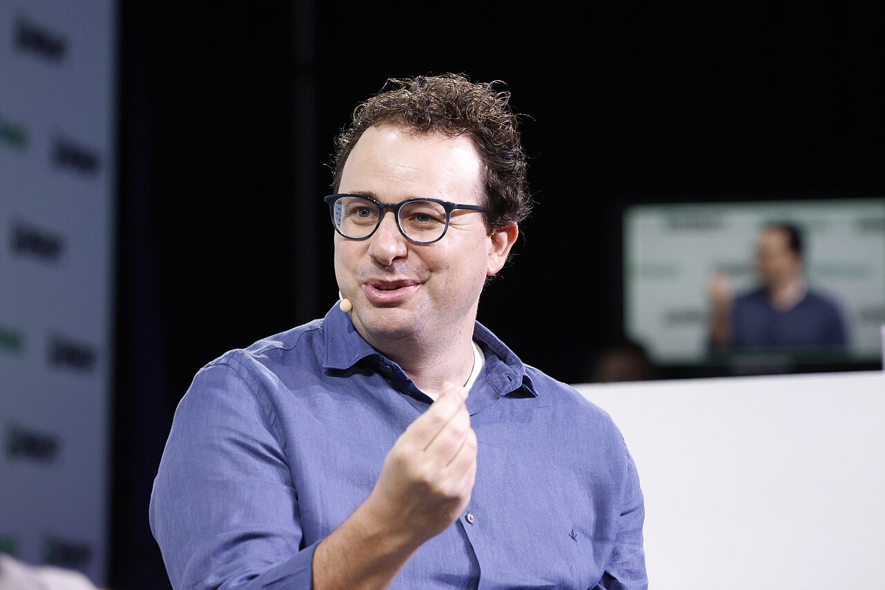 Dario_Amodei_at_TechCrunch_Disrupt_2023_01 Anthropic Co-Founder & CEO Dario Amodei speaking at TechCrunch Disrupt 2023, wearing glasses and a blue polo shirt, gesturing with his hand while presenting on stage with screens visible in the background