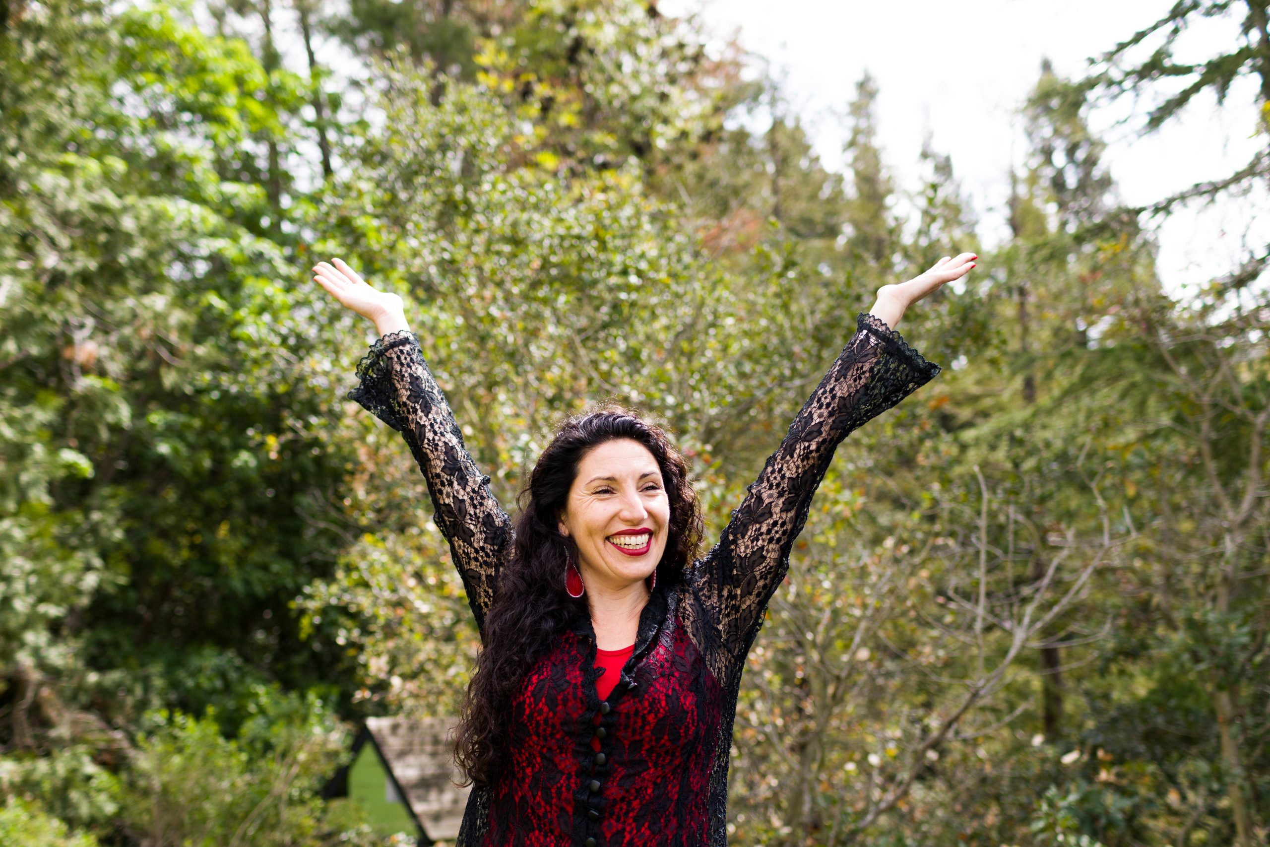 Maritza Schäfer with curly dark hair wearing a black lace top and red dress, arms raised joyfully in the air, smiling outdoors among trees in natural daylight