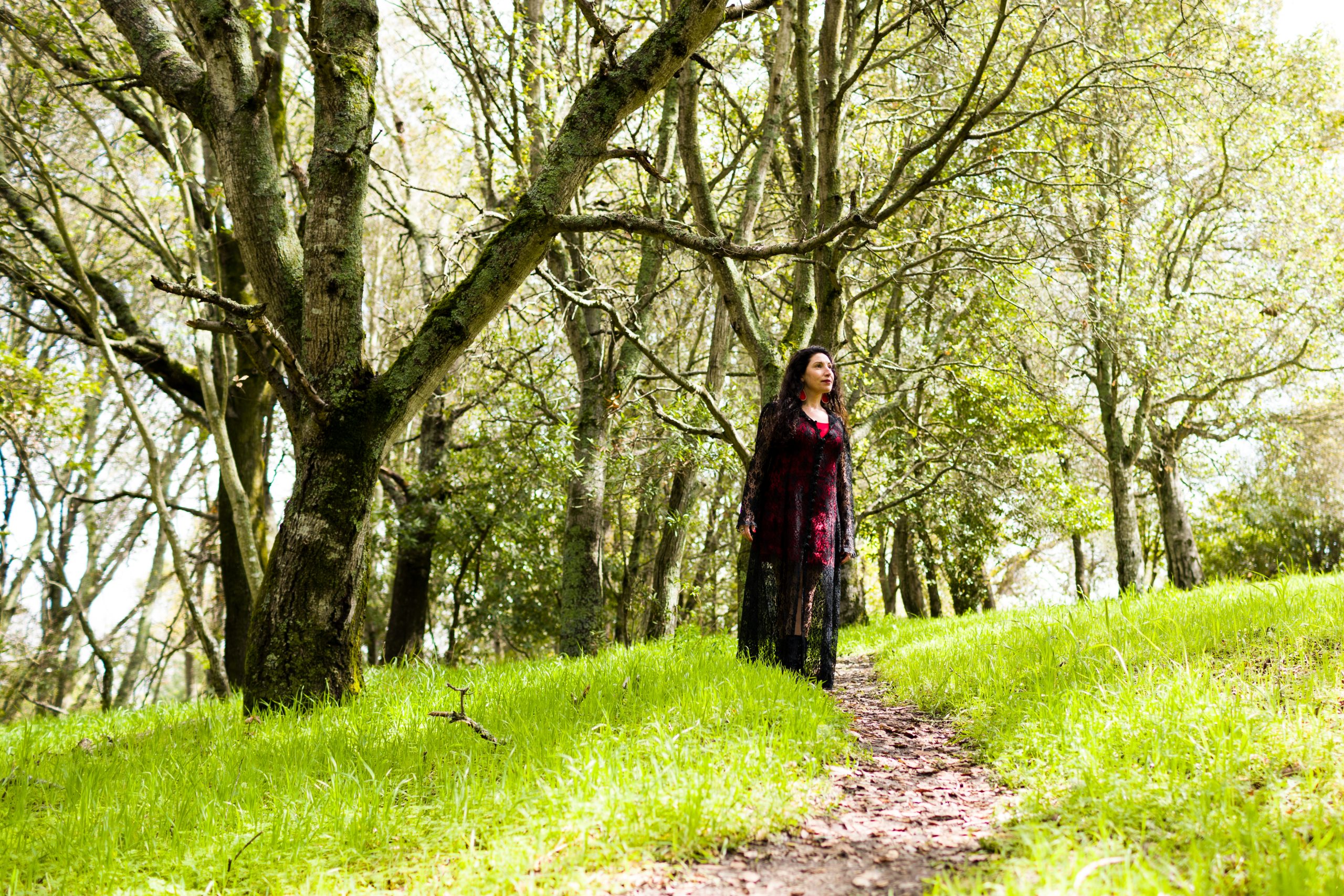 Maritza Schäfer standing on a dirt trail through a grove of moss-covered trees with bright green grass, wearing a dark red and black outfit