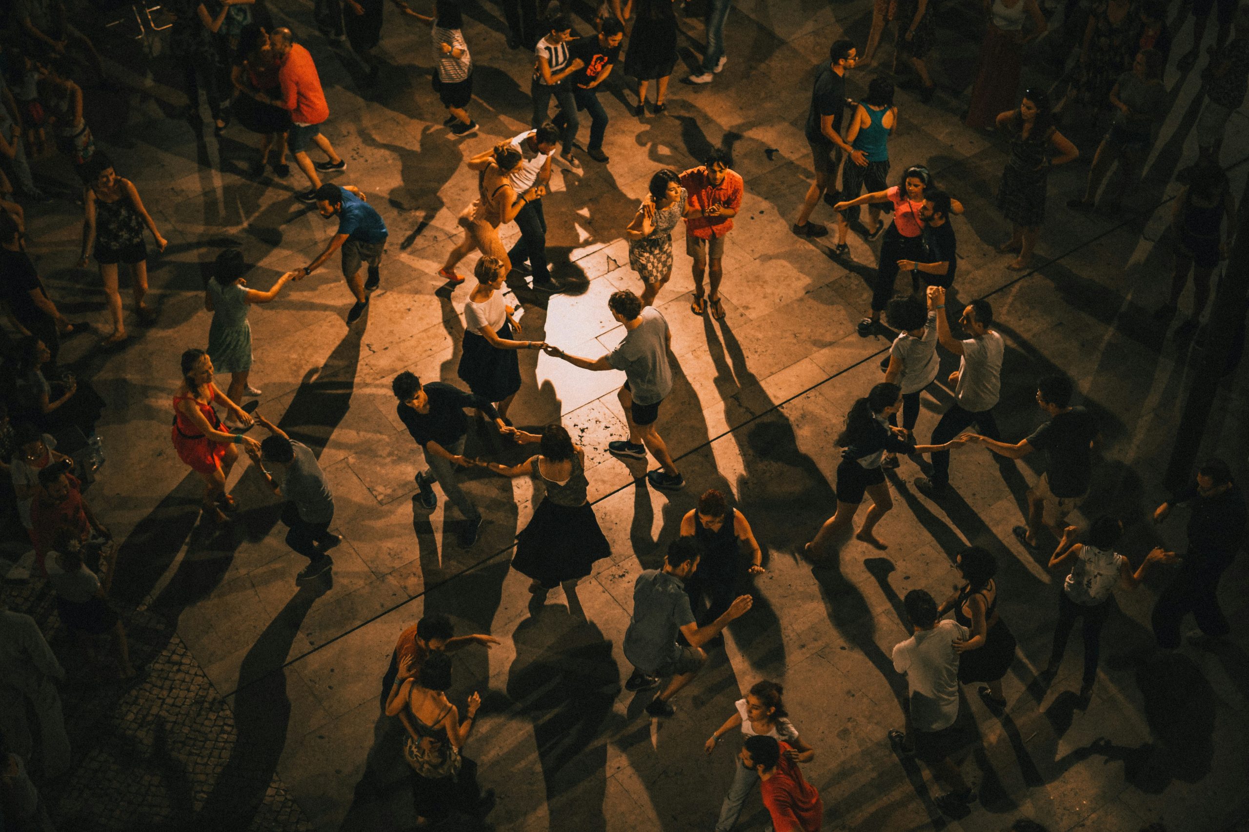 Overhead view of people dancing in a circle on a public plaza at dusk, with warm golden lighting casting long shadows on the pavement