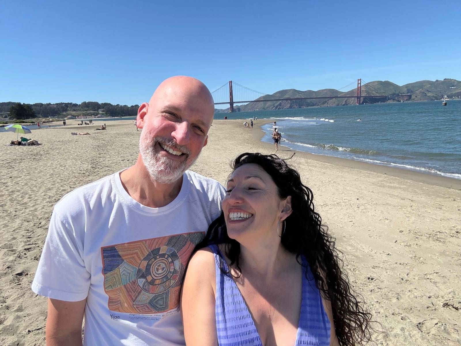 Maritza Schafer and her partner, Jeff, smiling on a sandy beach with the Golden Gate Bridge in the background on a sunny day