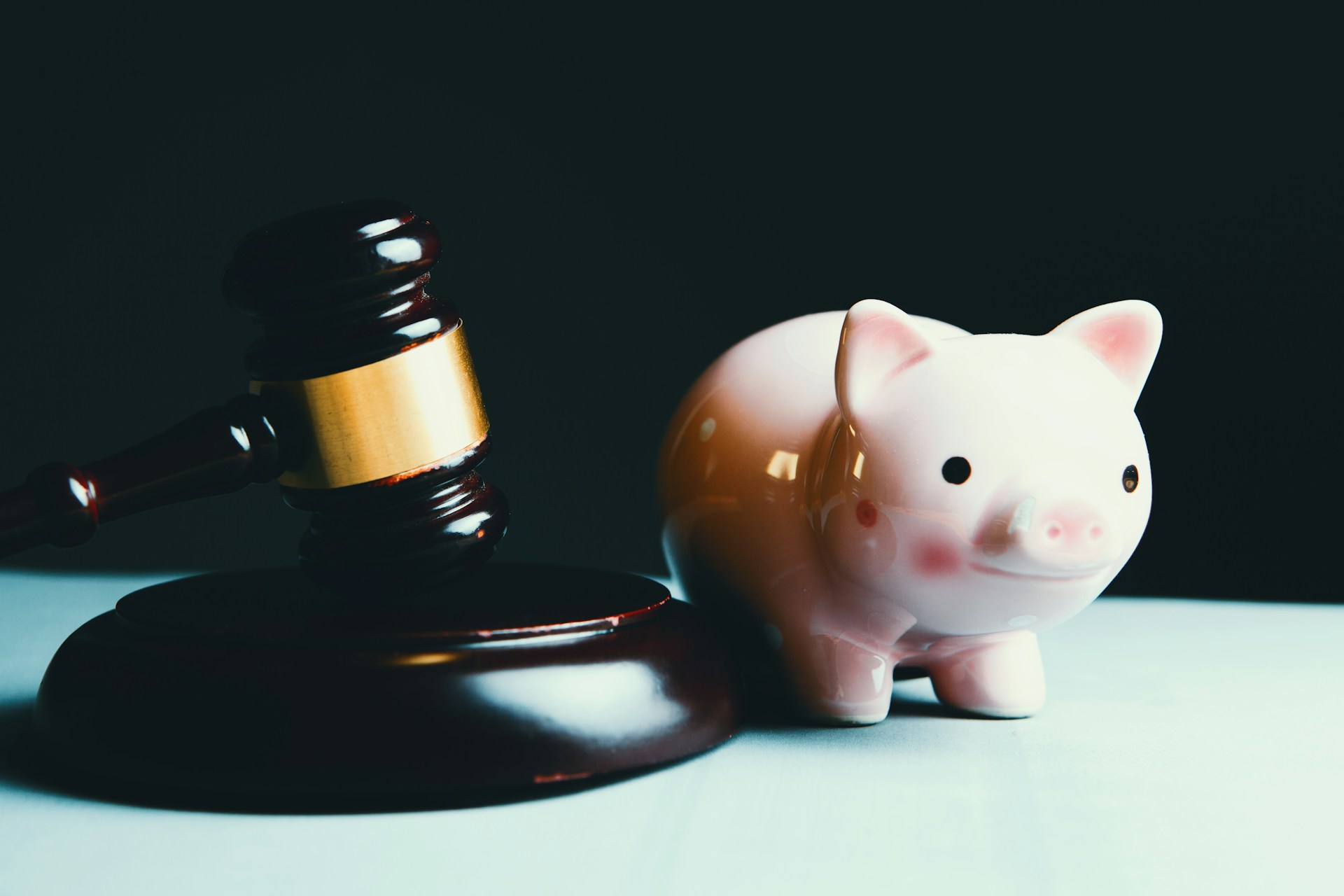 Pink ceramic piggy bank next to a wooden judge's gavel on a dark surface with dramatic lighting