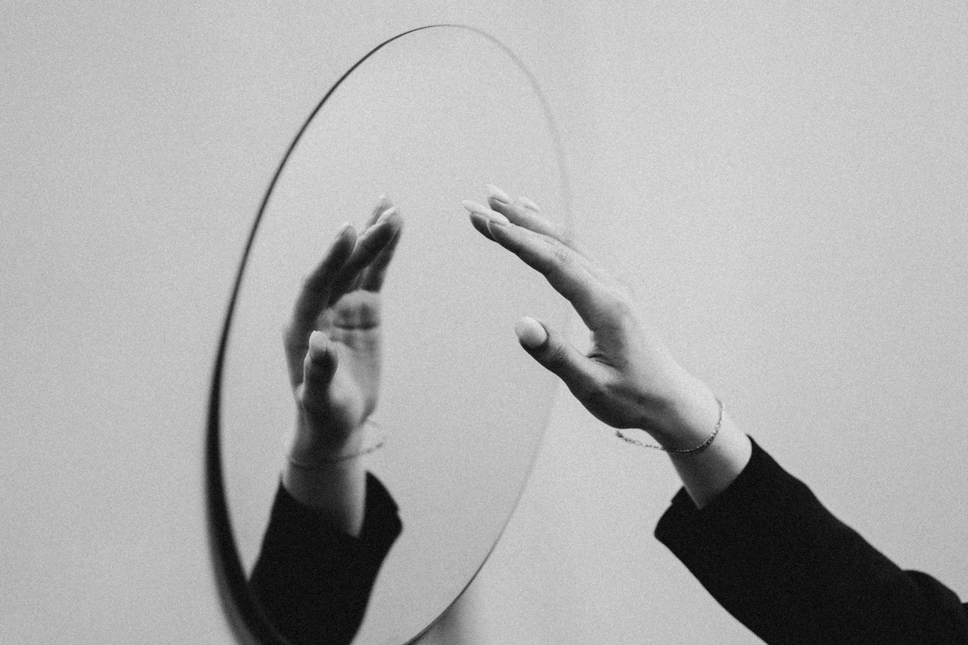 Black and white image of a hand reaching toward an oval mirror showing a blurred, distorted reflection against a plain background