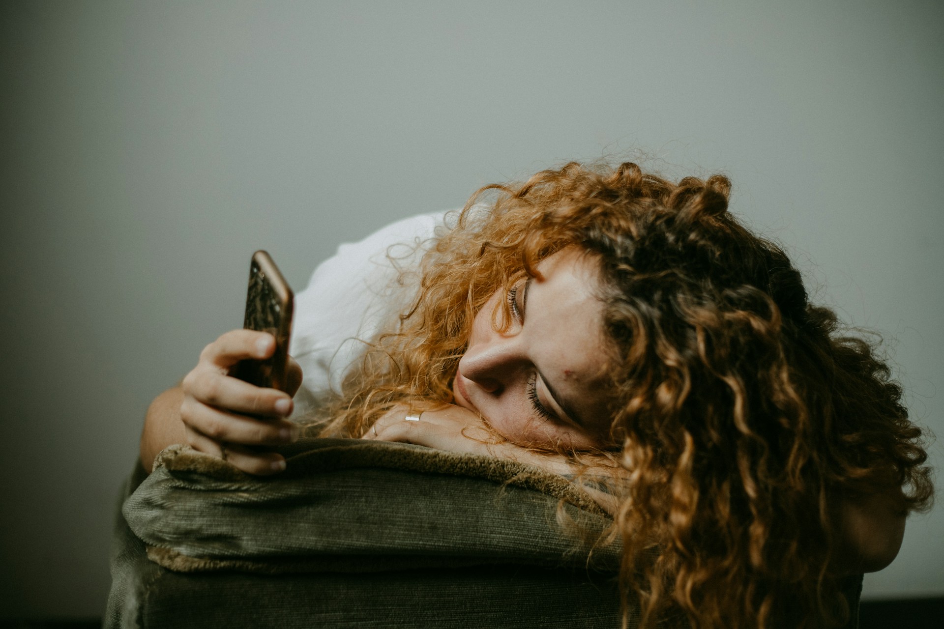 Woman with curly red hair lying down with eyes closed, holding a phone while resting her head on folded arms, appearing tired or distressed in soft natural lighting