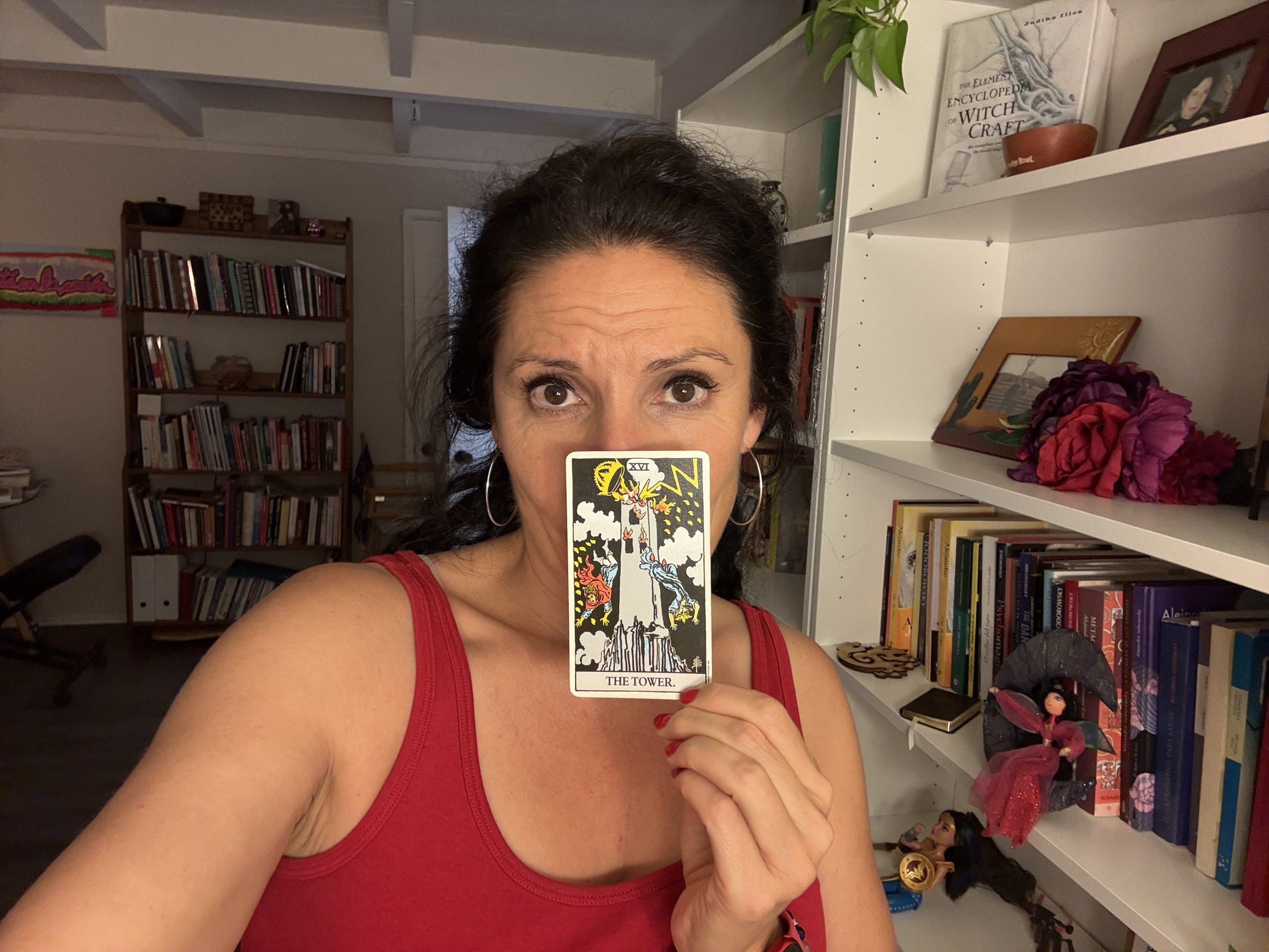 Maritza holding a Tower tarot card in front of her face, standing in front of a bookshelf filled with books and spiritual objects